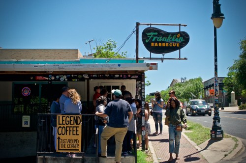 A crowd waits outside Franklin Barbecue on a sunny day, with a sign and nearby coffee shop visible.