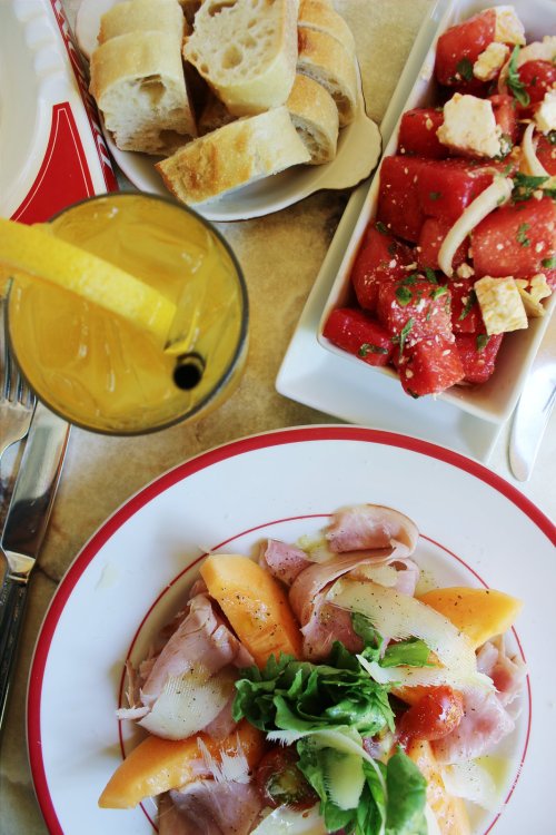 A colorful meal featuring a salad with ham and melon, a side of watermelon salad, bread, and a drink with a lemon slice.