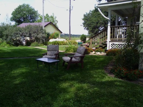 A peaceful garden scene with two chairs and a small table on green grass, near a house and trees.