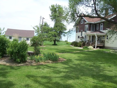 A large house with a porch and a smaller building nearby, surrounded by green grass and trees on a sunny day.