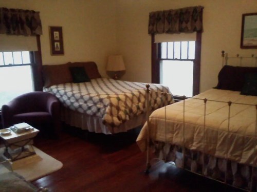 Cozy bedroom with two beds, a purple chair, and windows letting in natural light. Warm, inviting decor.