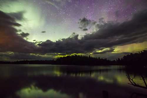 A serene lake reflects a starry sky with colorful auroras and clouds, creating a tranquil night scene.