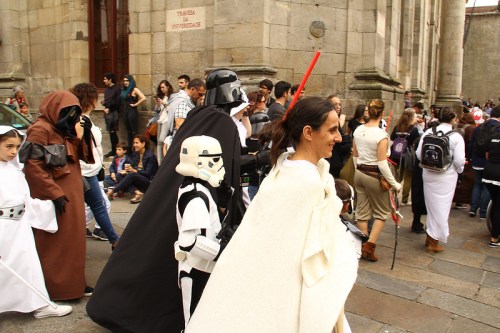 A group of people in Star Wars costumes, including Darth Vader and a stormtrooper, walking in a crowded outdoor setting.
