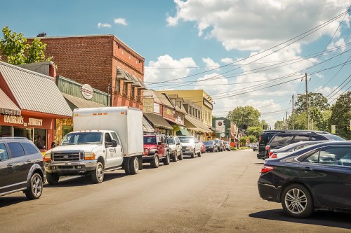 A sunny street scene with parked cars and shops lining both sides under a blue sky with fluffy clouds.