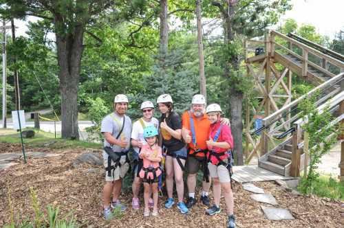 A group of six people, wearing helmets and harnesses, pose together outdoors near a zipline platform.