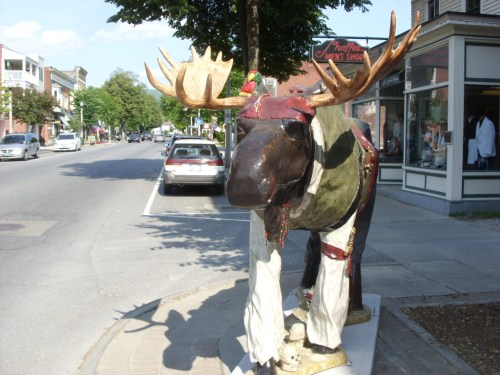 A colorful moose statue dressed in festive attire stands on a sidewalk near shops on a sunny street.