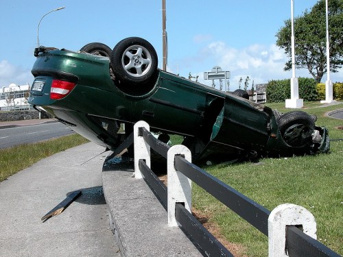 A green car is overturned on its side near a road, with debris scattered around and a clear sky in the background.