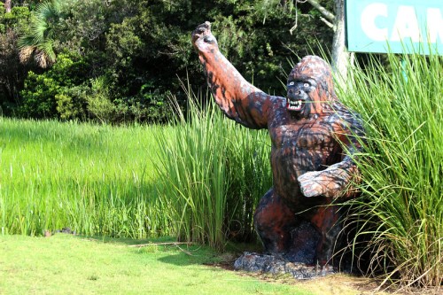 A large, colorful gorilla statue stands in tall grass, raising one arm in a playful pose against a green backdrop.