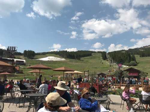 A busy outdoor area with people sitting at tables, mountains in the background, and a ski lift in the distance.