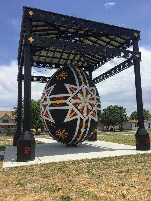 A large, decorative egg sculpture under a black metal canopy, surrounded by grass and buildings in the background.