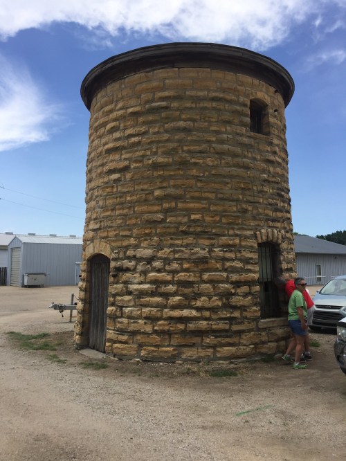 A round stone tower with a flat roof, surrounded by gravel and a few people nearby, under a blue sky.