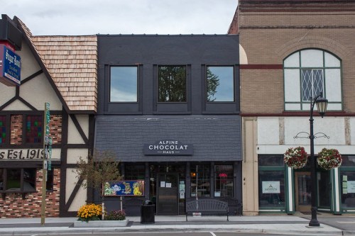 A storefront labeled "Alpine Chocolat Haus" with large windows, flanked by two other buildings, on a cloudy day.