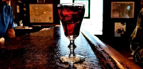 A glass of dark red beverage sits on a wooden bar, with a blurred background of a cozy pub setting.