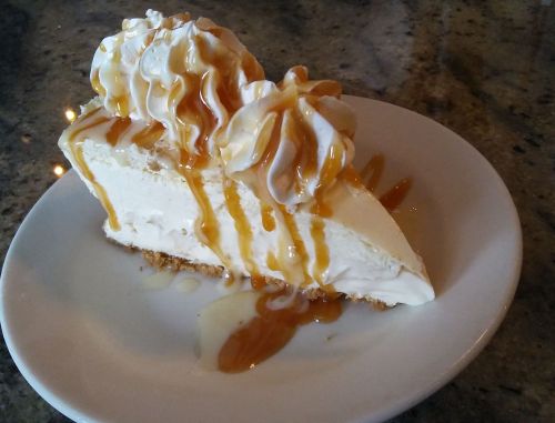 A slice of creamy cheesecake topped with whipped cream and drizzled with caramel sauce on a white plate.