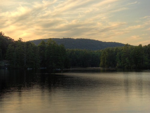 A serene lake surrounded by trees and mountains, reflecting a colorful sunset sky.