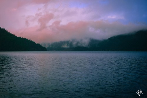 A serene lake surrounded by misty mountains under a colorful sky at dusk.