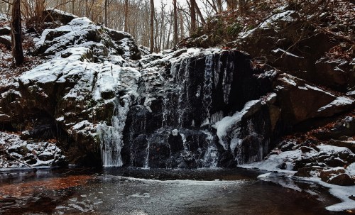 A serene waterfall cascades over rocks, surrounded by snow and bare trees in a tranquil forest setting.