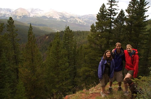 Three hikers pose on a rocky outcrop surrounded by tall trees, with mountains visible in the background.