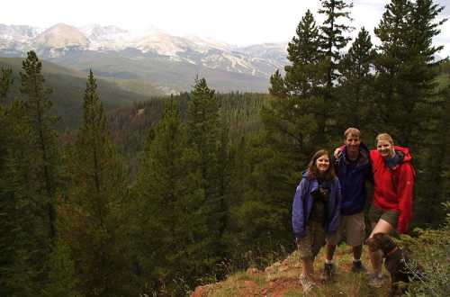 Three hikers pose on a rocky outcrop surrounded by tall trees, with mountains visible in the background.