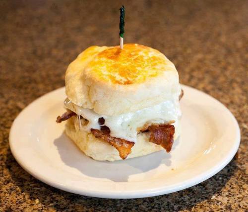 A biscuit sandwich with bacon and a fried egg, topped with a toothpick, on a white plate.
