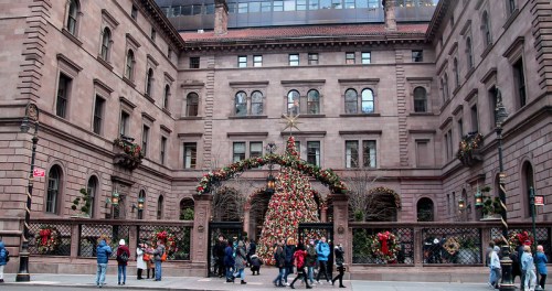 A festive courtyard decorated with a large Christmas tree and holiday decorations, bustling with people.
