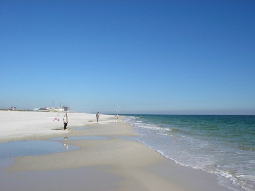 A serene beach scene with two people walking along the shore, clear blue sky, and gentle waves lapping at the sand.