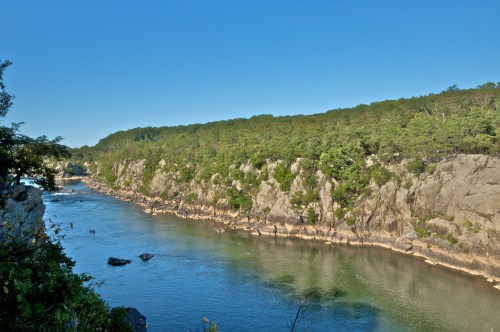 A serene river flows past rocky cliffs and lush greenery under a clear blue sky.