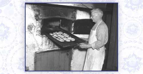 A baker in an apron removes a tray of bagels from a traditional oven.