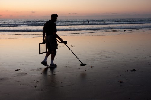 A person using a metal detector walks along a beach at sunset, with gentle waves in the background.