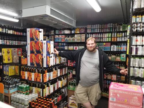 A man stands in a well-stocked beer cooler, surrounded by various beer boxes and cans on shelves.