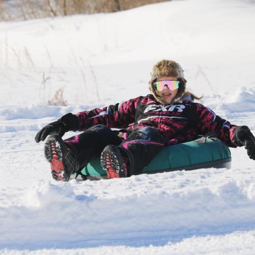 A person in a colorful winter outfit lies on a snow tube, enjoying a snowy landscape on a sunny day.