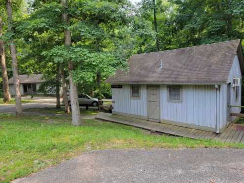 A small, light blue cabin surrounded by trees, with a wooden deck and a car parked nearby on a gravel road.