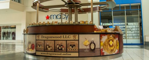 A retail kiosk in a mall featuring Dragonwood LLC products, with a Macy's store in the background.
