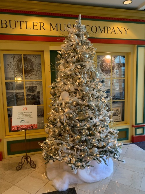 A beautifully decorated Christmas tree with white and silver ornaments, set in a festive lobby near a sign for "Eyesight Delight."