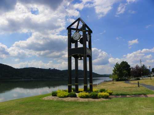 A tall clock tower beside a calm river, surrounded by green grass and trees under a partly cloudy sky.