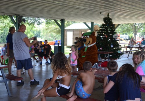 A group of children and adults gather under a pavilion, with costumed bears and a Christmas tree in the background.