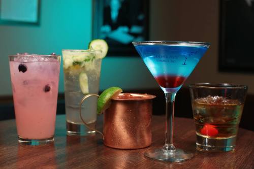 A variety of colorful cocktails on a wooden table, including a blue martini, a copper mug, and fruity drinks.