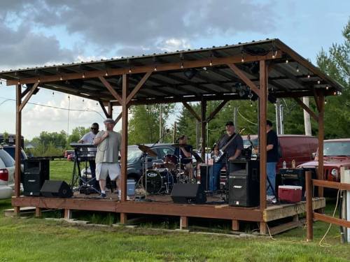 A band performs on a wooden stage outdoors, surrounded by greenery and parked cars under a cloudy sky.