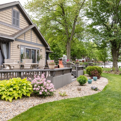 A cozy lakeside home with a deck, surrounded by lush greenery and colorful flower beds.