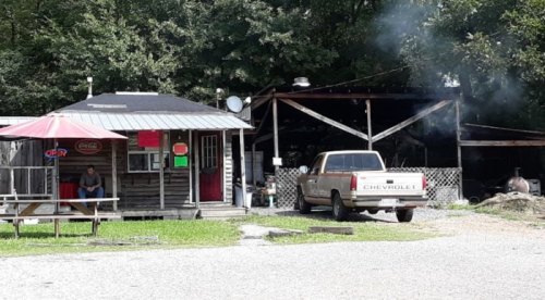 A rustic roadside stand with a picnic table, a truck parked nearby, and smoke rising from a grill in the background.