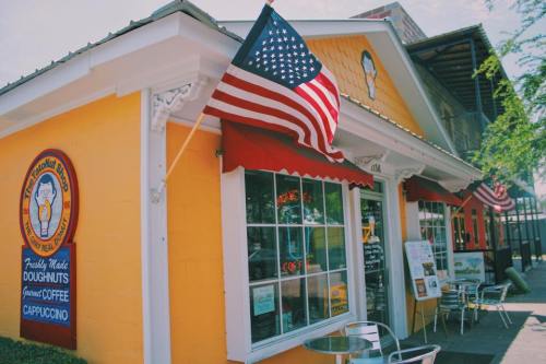 A bright yellow café with an American flag, featuring a sign for doughnuts and coffee, and outdoor seating.
