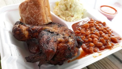 A takeout container with grilled chicken, baked beans, coleslaw, and a bread roll, served with a side of sauce.