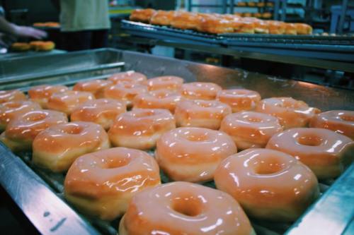 Freshly glazed donuts arranged on a tray, with more donuts in the background.