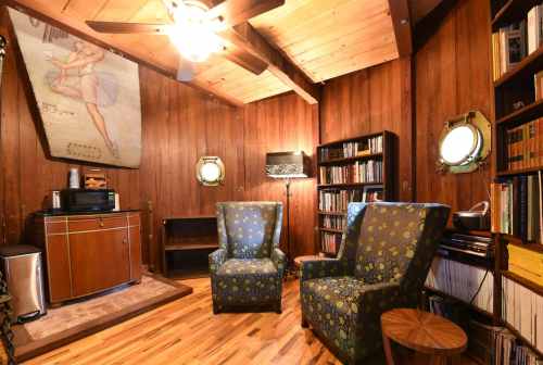 Cozy wooden room with two patterned chairs, a vintage poster, bookshelves, and a small kitchenette area.