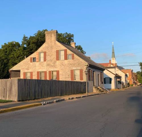 A historic stone house with red shutters, surrounded by trees, on a quiet street with a church steeple in the background.