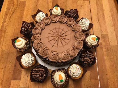 A chocolate cake with a decorative frosting design, surrounded by assorted chocolate and vanilla cupcakes.
