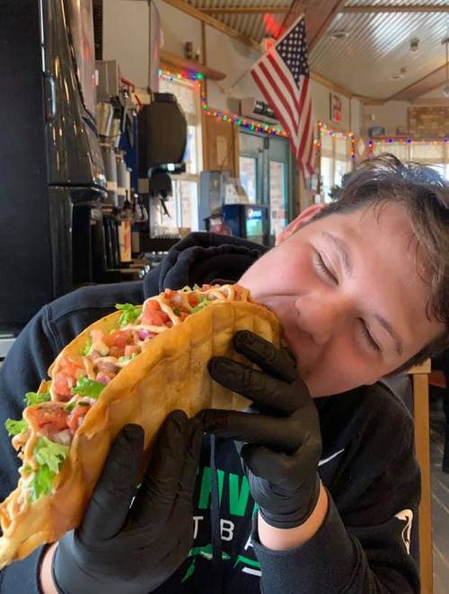 A person wearing gloves prepares to take a big bite of a loaded taco in a cozy restaurant setting.