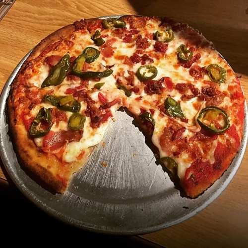 A pizza with bacon, jalapeños, and melted cheese, partially sliced on a metal tray.