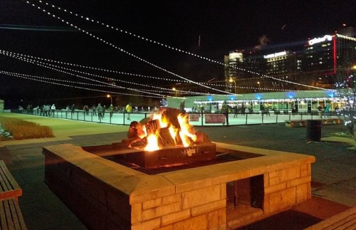 A cozy fire pit surrounded by string lights at night, with people in the background enjoying the atmosphere.