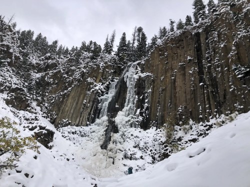 A frozen waterfall cascading down a rocky cliff, surrounded by snow-covered trees and a snowy landscape.